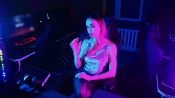 Концепция игры - молодая женщина с татуировками сидит в кресле в неоновом игровом клубе и высасывает леопарда — стоковое видео