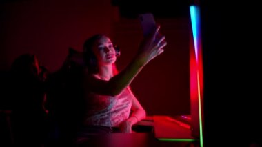 Neon oyun kulübünde oturan ve telefonuyla selfie çeken dövmeli genç bir kadın.