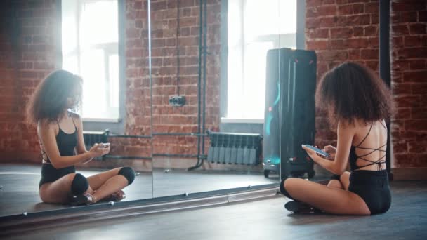 Strip dançando no estúdio loft - jovem dançarina encaracolada sentada em seu telefone na frente do espelho — Vídeo de Stock