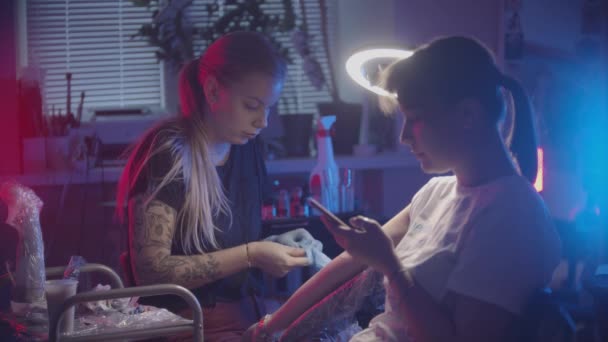 Sesja tatuażowa - mistrz tatuażu zakładający rękawiczki i jej klient sprawdzający jej telefon — Wideo stockowe