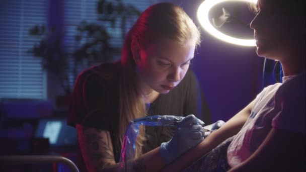 Sesja tatuażowa - zrobienie czarnego abstrakcyjnego tatuażu na ramieniu przez panią mistrz w ciemnym studiu z kolorowym oświetleniem — Wideo stockowe