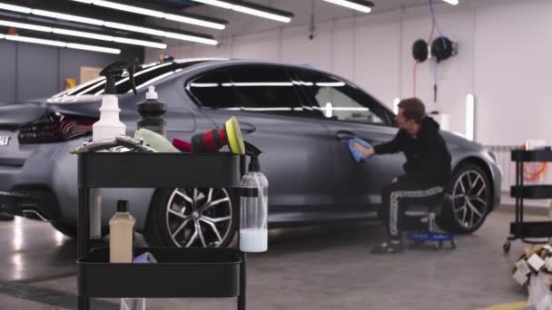 Automatisk detalj salong - man arbetare sitter nära den ljusgrå bilen och polera ytorna - rengöringsmedel på förgrunden — Stockvideo