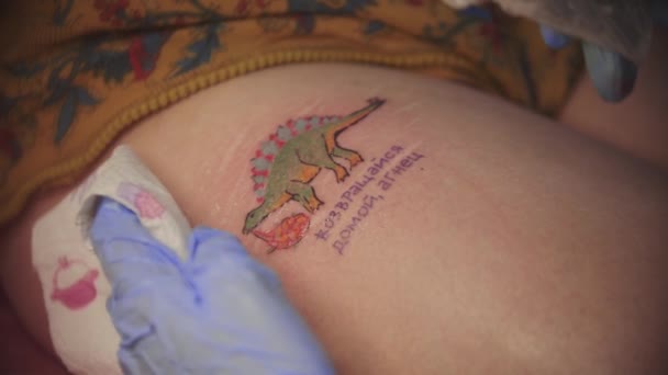 Una mujer haciéndose un tatuaje de dinosaurio colorido con palabras rusas — Vídeos de Stock
