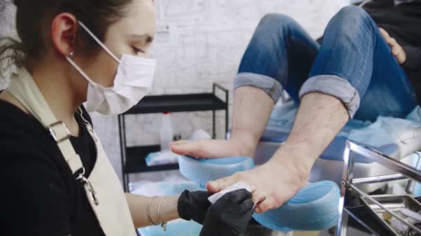 Мастер по педикюру вытирает ногтевую пыль с ног ее клиента-мужчины — стоковое видео