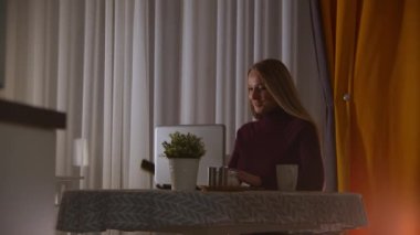 Evde çalışan sarışın bir iş kadını bilgisayarını kullanıyor ve telefonunu kontrol ediyor.