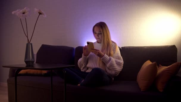 Een blonde vrouw zit op een bank en scrollt haar feed aan de telefoon — Stockvideo