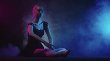 Siyah elbiseli genç kadın balerin yerde oturup esneme hareketleri yapıyor.