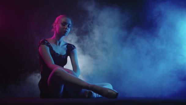 Junge Ballerina in schwarzem Kleid sitzt auf dem Boden im schwarzen rauchigen Studio und macht Stretching — Stockvideo