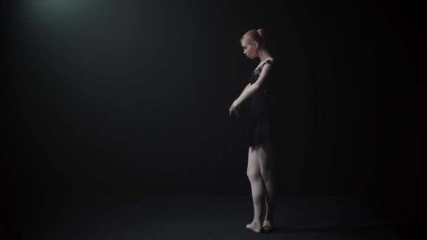 Молодая грациозная балерина в черном платье медленно танцует в черной студии — стоковое видео