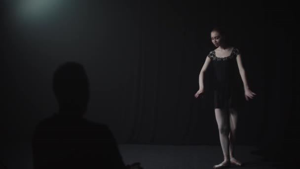 Joven bailarina elegante en vestido negro bailando lentamente en el estudio negro - el público observándola bailar — Vídeo de stock