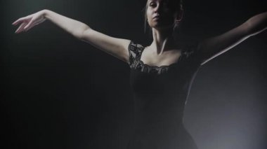 Siyah elbiseli genç kadın balerin dumanlı sahnede yavaşça dönüyor.