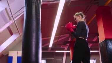 Genç bir kadın yumruklarını kırmızı spor salonundaki kum torbasında eğitiyor.