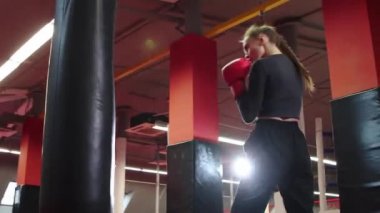 Uzun saçlı genç bir kadın kırmızı spor salonundaki kum torbasında yumruklarını eğitiyor.