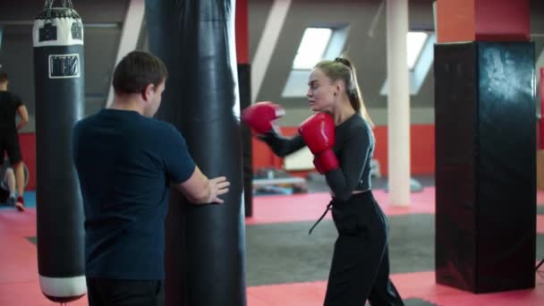 Entraînement à la boxe - une jeune femme séduisante en vêtements de sport frappe le sac et son entraîneur debout derrière — Video