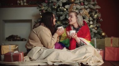Genç lezbiyen kadınlar Noel ağacının altında oturup kakao içerler.