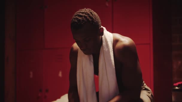 Ciemnoskóry młodzieniec siedzi w szatni z ręcznikiem na szyi. — Wideo stockowe