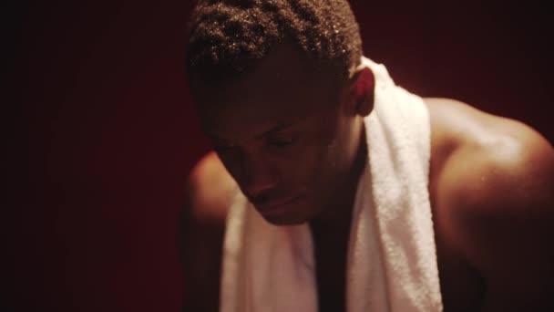 Jeune homme noir assis dans un vestiaire avec une serviette autour du cou - lumière reflétant différemment — Video