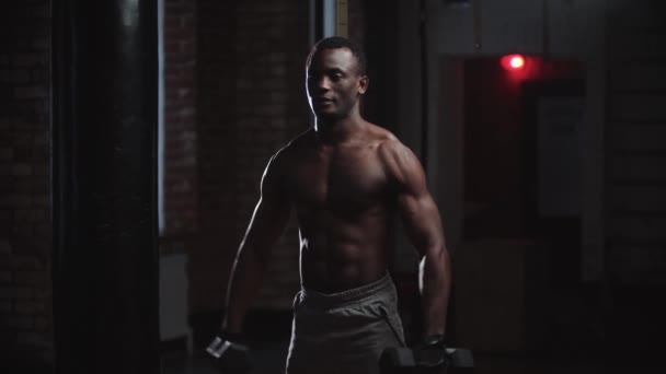 Um homem negro bombeando as mãos músculos com halteres no ginásio escuro — Vídeo de Stock
