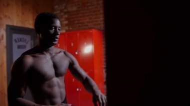 Afro-Amerikan atletik çıplak genç adam soyunma odasındaki dolabını açıyor.