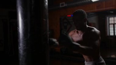 Siyah atletik üstsüz genç adam boksör eğitimi. Spor salonunda çabalayarak kum torbasını yumrukluyor.