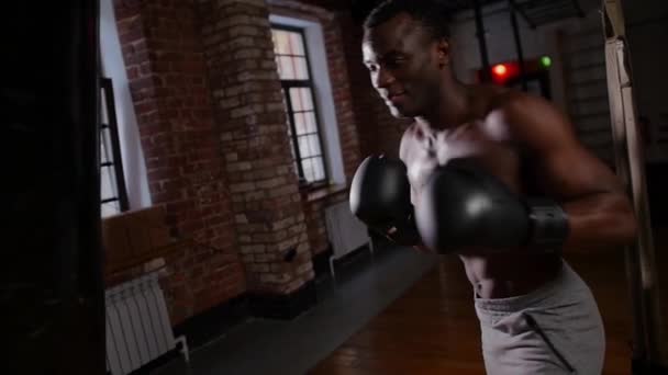 Afro-americano atlético sem camisa jovem boxeador treinamento soco no saco de perfuração com um esforço no ginásio — Vídeo de Stock