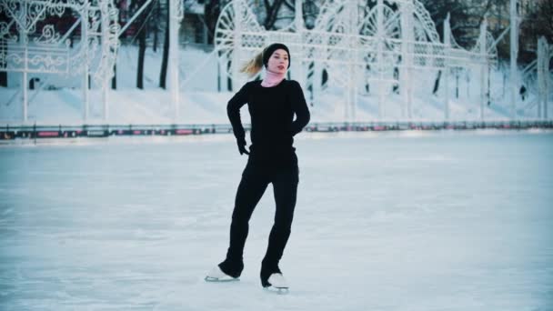 Mujer rubia joven en ropa negra con alta cola de caballo patinaje artístico en la pista de hielo pública por la mañana — Vídeo de stock