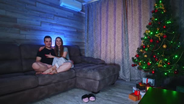 Pasangan muda duduk di sofa dan menonton TV sambil saling berpelukan — Stok Video