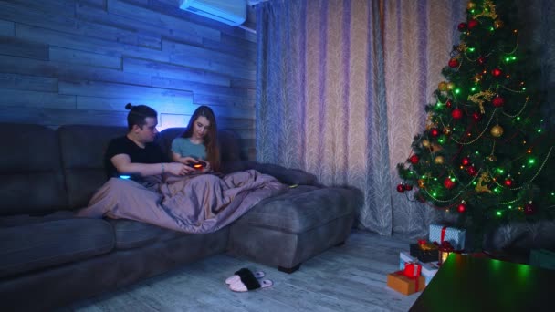 Pasangan muda duduk di sofa di bawah selimut dan bermain game di TV pacar mengajarkan pacarnya bagaimana mengendalikan joystick — Stok Video