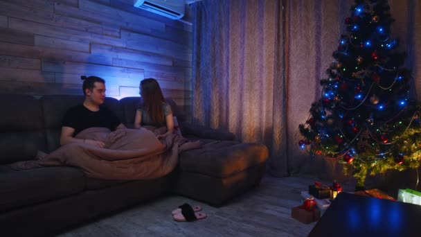 Mladý pár sedí na gauči pod přikrývkou - žena dává svému příteli vánoční dárek — Stock video