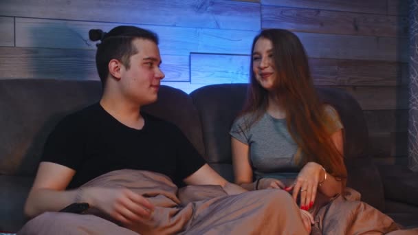 Mladý roztomilý pár sedí na gauči pod dekou - muž dává své přítelkyni vánoční dárek — Stock video