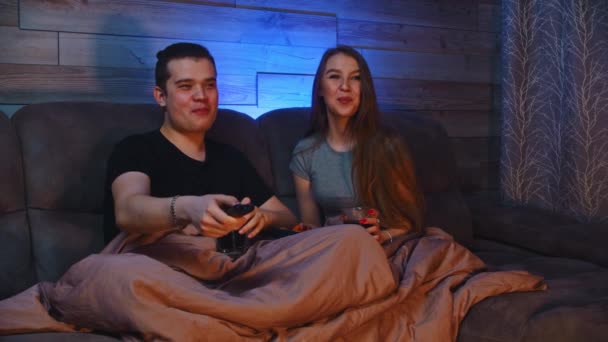 Jovem casal feliz sentado em um sofá sob cobertor e assistindo TV e comer lanches — Vídeo de Stock