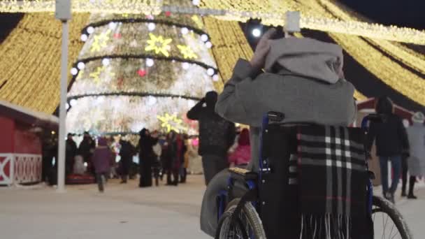 Una giovane donna sulla sedia a rotelle guarda un albero di Natale luminoso e folle di persone — Video Stock