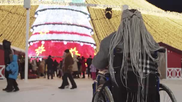 Seorang wanita muda dengan rambut gimbal abu-abu di kursi roda melihat pohon Natal yang cerah dan kerumunan orang — Stok Video