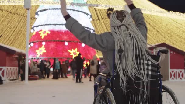 Tekerlekli sandalyedeki gri saçlı genç bir kadın parlak bir Noel ağacına ve kalabalığa bakıyor ve hafifçe dans ediyor. — Stok video