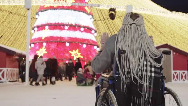 Una joven con rastas grises en silla de ruedas mira un árbol de Navidad y multitudes de personas y bailando ligeramente — Vídeos de Stock