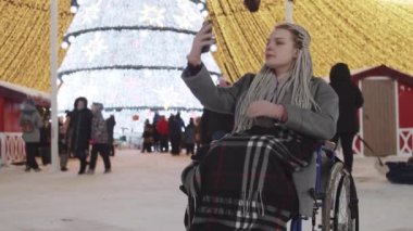 Noel partisinde tekerlekli sandalyedeki genç bir kadın sokaklarda fotoğraf çekiyor ve rastalarını tamir ediyor.