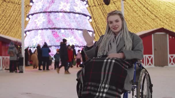 Jeune femme en fauteuil roulant à la fête de Noël dans la rue - souriant et agitant la main — Video