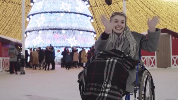 Joven mujer positiva en silla de ruedas en la fiesta de Navidad en las calles sonriendo y agitando la mano — Vídeos de Stock