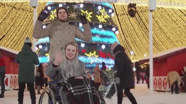 Mujer joven en silla de ruedas y su novio en la fiesta de Navidad en las calles saludando con las manos a la cámara — Vídeos de Stock