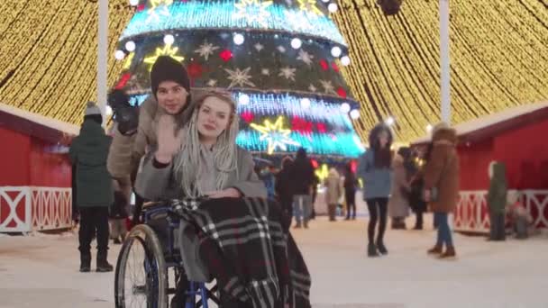 Χαμογελαστή νεαρή γυναίκα σε αναπηρική καρέκλα και ο φίλος της στο χριστουγεννιάτικο πάρτι στους δρόμους - χαιρετώντας την κάμερα — Αρχείο Βίντεο