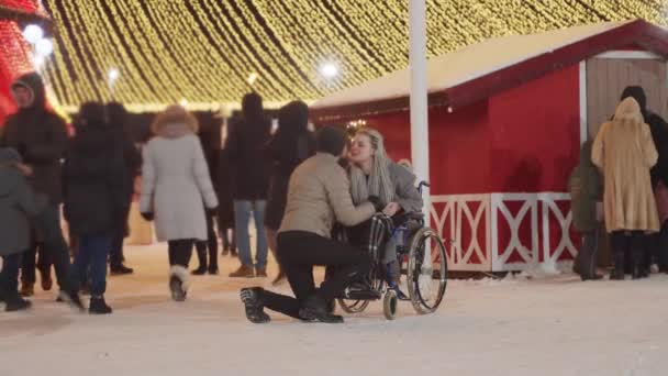 Glimlachende jonge vrouw in een rolstoel en haar vriendje op kerstfeest op straat - zoenen — Stockvideo