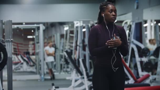 Mujer negra insegura de pie en el gimnasio con auriculares — Vídeos de Stock