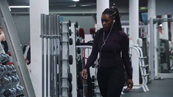 Czarna kobieta w słuchawkach spaceruje po sali gimnastycznej i bierze hantle — Wideo stockowe
