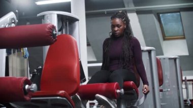 Spor salonunda egzersiz aletleriyle çalışan Afrikalı Amerikalı bir kadın bacak kaslarını çalıştırıyor.
