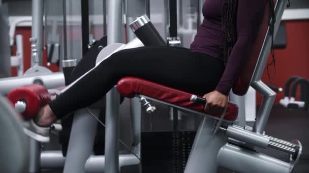 Een zwarte vrouw met vlechten uit te werken in een sportschool op een been fitnessapparaat — Stockvideo