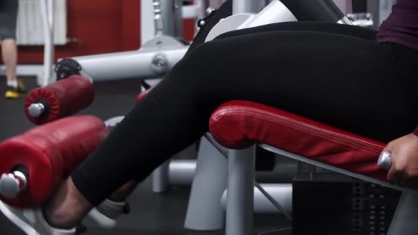 Uma mulher negra com tranças treinando as pernas em um equipamento de exercício e mastigando uma gengiva — Vídeo de Stock