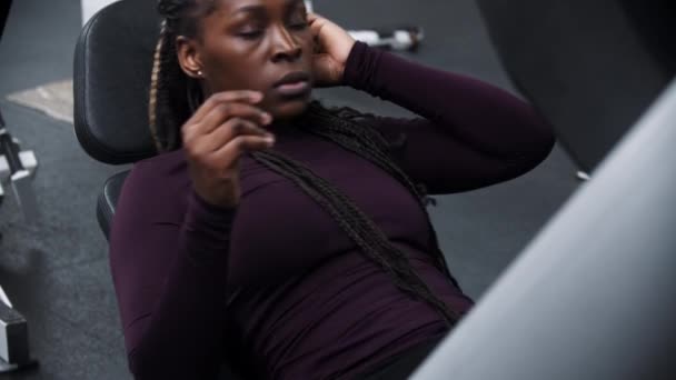 A black woman with braids sits down on a exercise equipment for legs — Stock Video