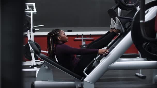 En svart kvinna med flätor sätter sig på ett träningsredskap för ben och börjar träna — Stockvideo
