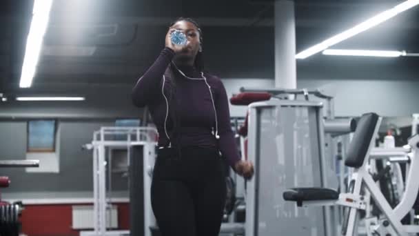 Afroameričanka ve sluchátkách chodící v tělocvičně a pitné vodě — Stock video