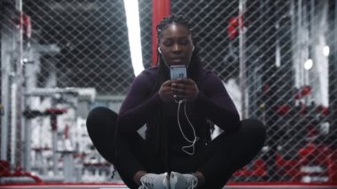 Gülümseyen Afro-Amerikalı kadın spor salonunda yoga minderinde oturuyor telefonunu kullanıyor ve müzik dinliyor.